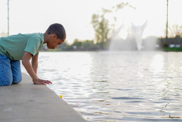 A young child kneels at the edge of a fountain in Bentonville, Arkansas. The child appears to be intently watching a small yellow object, possibly a toy duck, floating on the water's surface.