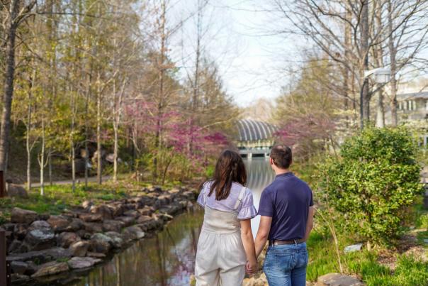 A couple holds hands while admiring the view over a rocky stream at Crystal Bridges Museum of American Art, surrounded by trees showing the colors of spring.