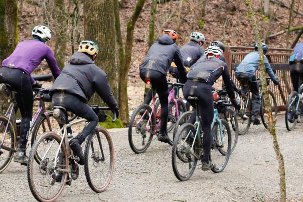 A group of cyclists in active gear rides along a gravel path through a wooded area, approaching a gate. Trees surround the trail.