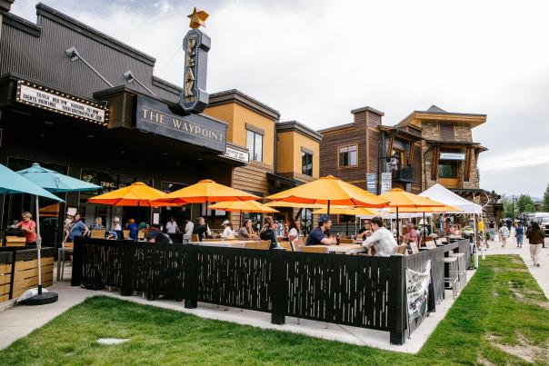 Outdoor dining scene at The Waypoint, featuring vibrant orange umbrellas and a lively crowd enjoying meals at wooden tables. Nearby buildings add charm.