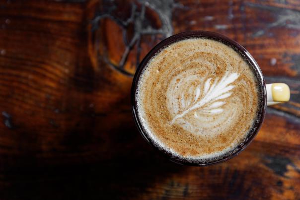 A close-up of a latte in a dark ceramic cup, featuring intricate latte art resembling a leaf, set on a rustic wooden table.