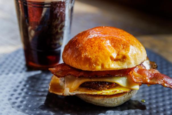 A close-up of a breakfast sandiwch with bacon and cheese, served on a shiny bun, alongside a glass of cold brew on a textured plate.