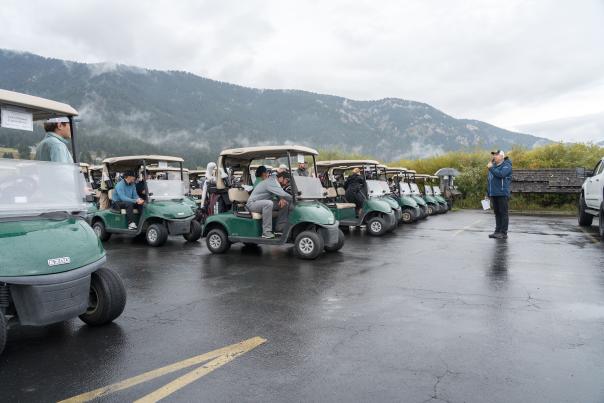 A row of golf carts parked in a lot, with a group of people gathered around a guide speaking. Mountains and cloudy skies in the background.