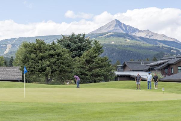 A scenic golf course with a putting green, surrounded by trees and mountains under a partly cloudy sky. Three golfers are on the green.