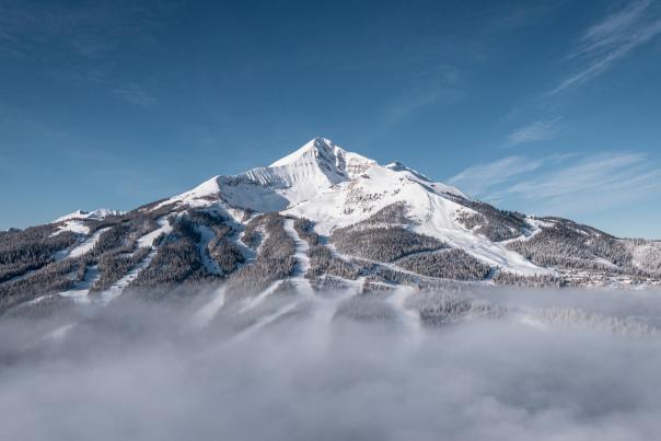 Snow-covered mountain peak rises above a blanket of clouds, surrounded by evergreen forests and ski trails under a clear blue sky.