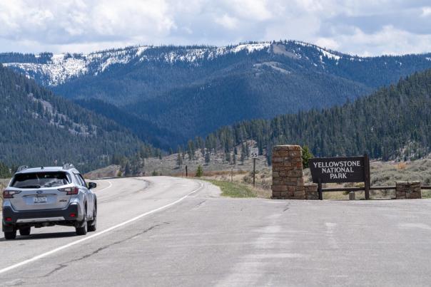 A car approaches the entrance to Yellowstone National Park, with snow-capped mountains and lush greenery in the background. A sign reads "Yellowstone National Park."