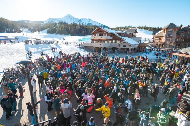A bustling ski resort scene with a large crowd in colorful winter attire, surrounded by snow-covered mountains and lodges.