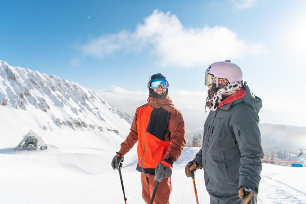 Two skiers stand on a snowy slope, surrounded by mountains under a clear blue sky. They wear winter gear and helmets, smiling.