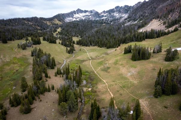 Aerial view of a lush green valley surrounded by rugged mountains, with winding trails and a small stream flowing through the landscape.