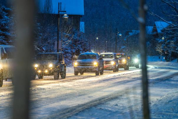 A photo of cars lined up on a snowing street at dusk with their headlights on.