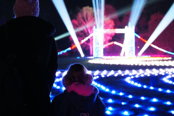 A mother and small child stand with their backs to the camera looking at a small, lit up London Bridge. There are roaming spotlights & glowing fairylights on the floor in all different colours. The trees off in the distance are also lit up in blue and pink.