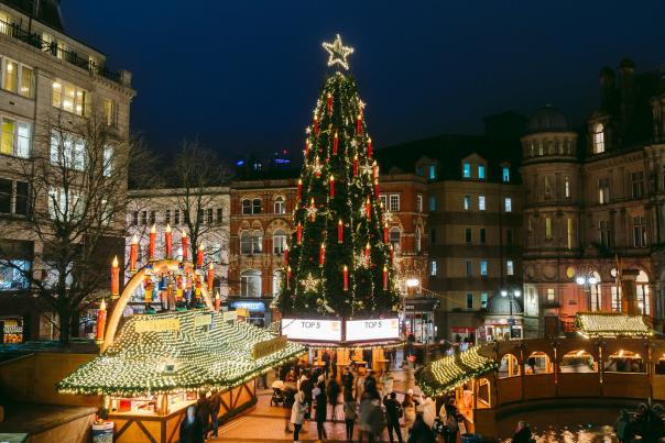 A tall Christmas tree stands in a square in Birmingham at night, the lights are lit and people are gathered round its base at Christmas market stalls