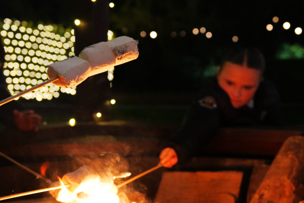 Two little girls roasting marshmallows over a fire pit. In the foreground is a stick with three roasted marshmallows. Blurred in the background are the two children. In the distance in bokeh and twinkling lights.