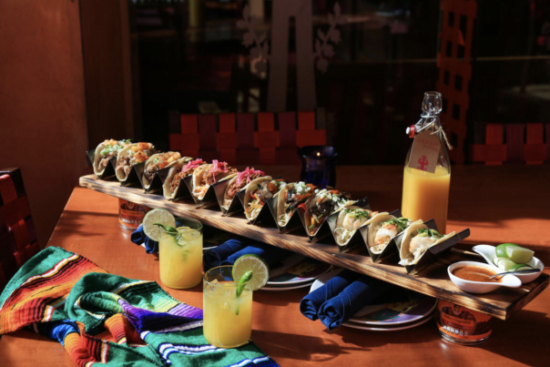 Long row of tacos on a board with two citrusy-looking drinks at Rosa Mexicano