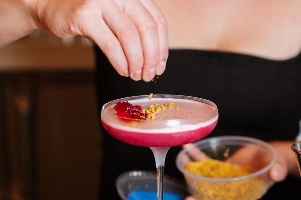 A mixologist tops a bright pink cocktail off with a pinch of yellow bee pollen