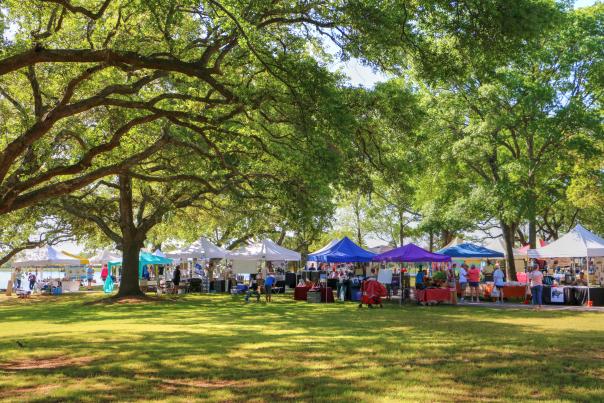 Visitors explore a vibrant outdoor market in Sunset Beach, NC, with colorful vendor tents under shady trees on a bright, sunny day.