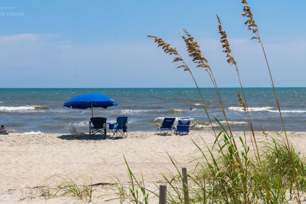 a view of a beach with an umbrella and chairs from behind a line of sea oats