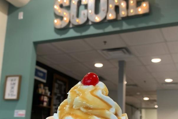 Scoop and Score Sundae in a pink and white polka dot cup with caramel sauce on top and a maraschino cherry