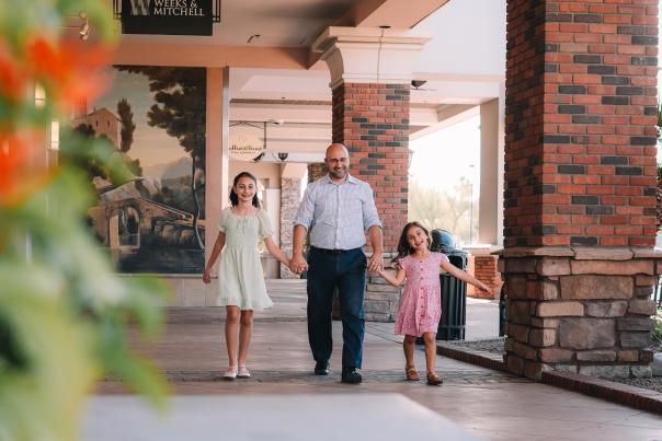 Two young brunette girls in spring-like dresses standing on either side of their father, wearing a light button up shirt and dress pants, holding his hands walking down the sidewalk in front of stone storefronts smiling.