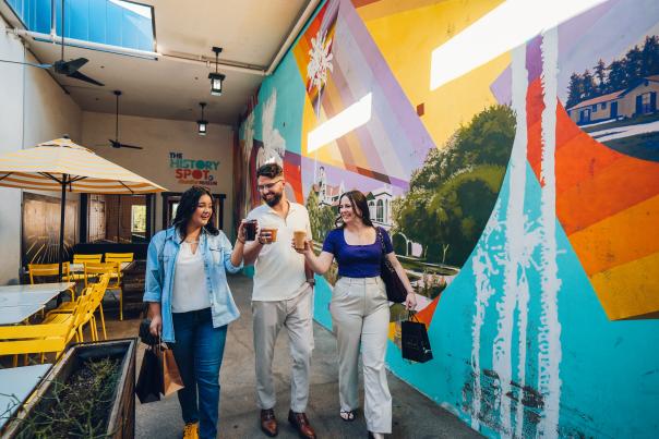 Group of 2 ladies and 1 gentleman walking past the colorful "Monuments to Chandler" mural, cheering with their drinks.