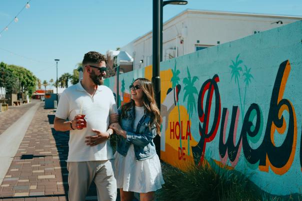 Couple holding drinks and smiling walking down street with colorful mural along wall.