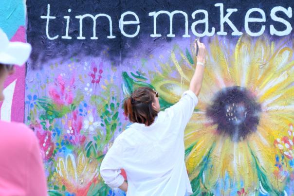 A person in pink with their back to us watches an artist painting bold sunflowers on a purple background