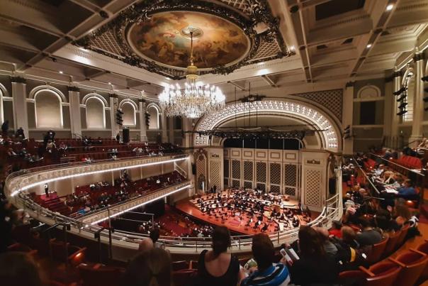 Inside an amphitheater from the balcony. White lights frame the stage and line the edges of the balconies. A small orchestra is playing to an audience