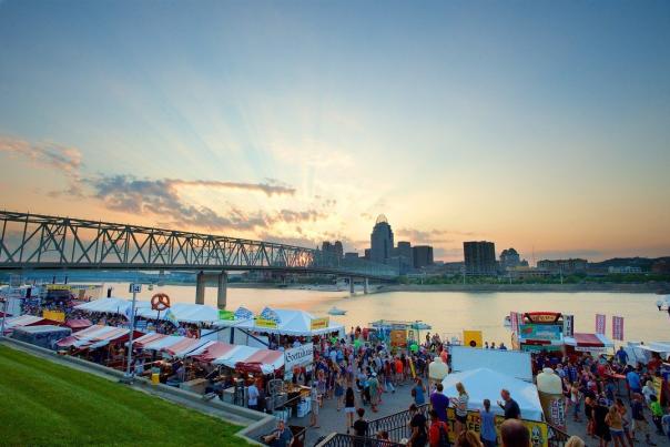 Image is of the Newport Festival Park with people walking around and the skyline of Cincinnati in the background.