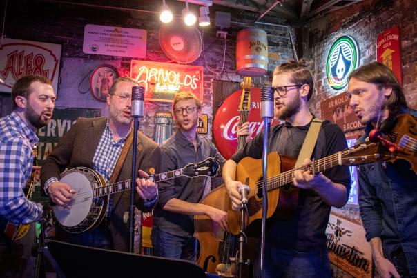 5 musicians gather around a couple of microphones. There's a banjo, guitar, upright bass, and violin visible. Behind them is a wall full of signs for beverages. Neon letters spell out Arnold's Est. 1861