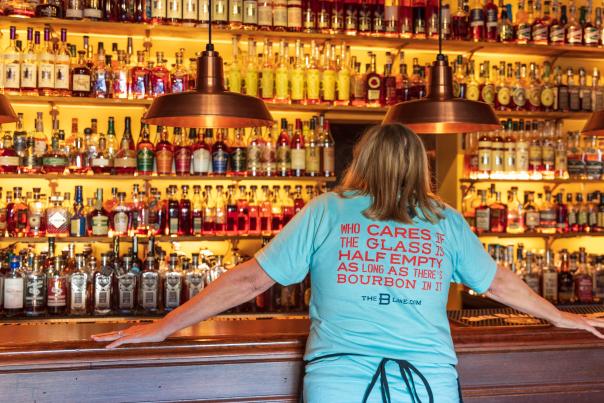 A woman in a light blue shirt stands in front of seemingly hundrands of bourbon botttles. Her shirt reads, "Who cares if the glass is half empty as long as there's bourbon in it" The B-Line