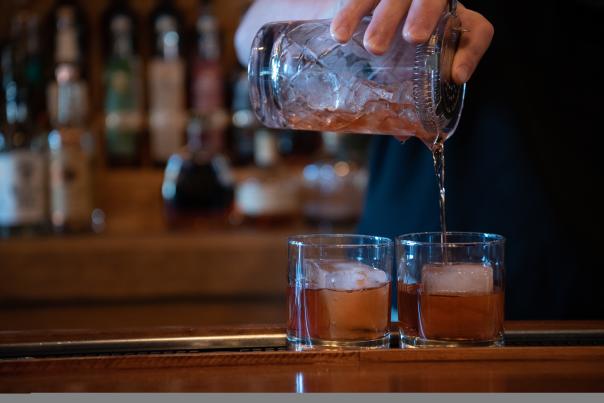 A close up of two drinks being poured over large, square ice in rocks glasses.