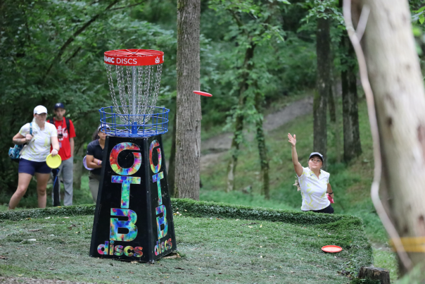 A woman in a white shirt throws a disc into a disc golf basket