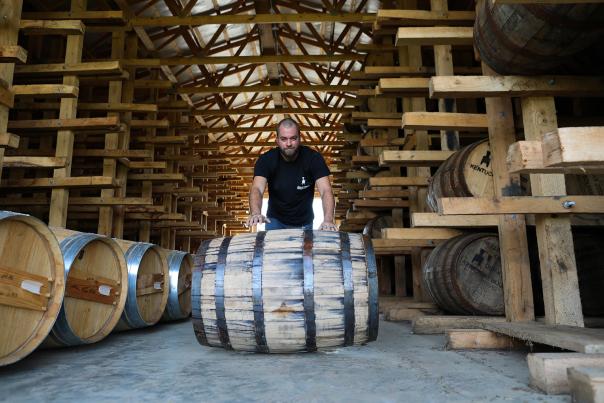 A person in a black t-shirt with a shaved head pushes a large bourbon barrel at Neeley Family Distillery's rickhouse