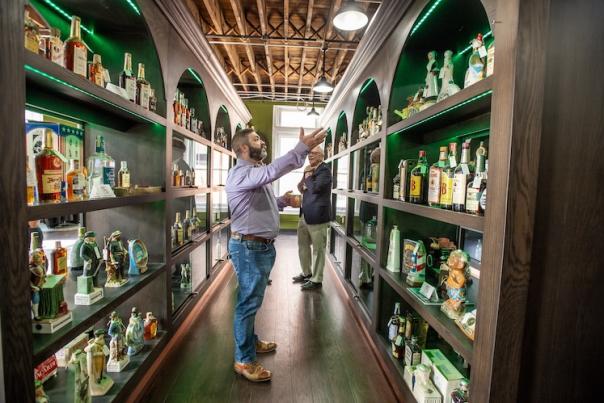 Two men stand looking at bookshelves filled with bottles of vintage spirits