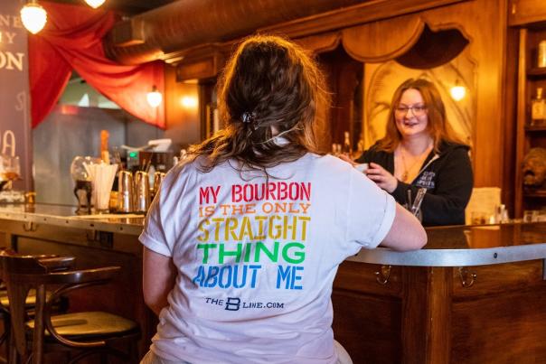 A woman wearing a t-shirt that reads in rainbow letters "My bourbon is the only straight thing about me" seated at the bar at Second Sight Spirits with a woman bartender offering her a drink.