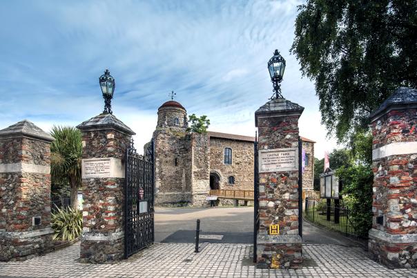 Colchester Castle photographed from outside the Castle Park gates. The Gates are open and four chunky stone pillars with lamps on top sit in the foreground.