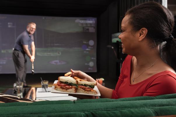 Woman enjoys sandwich while watching her husband play a virtual game of golf at the Main Course