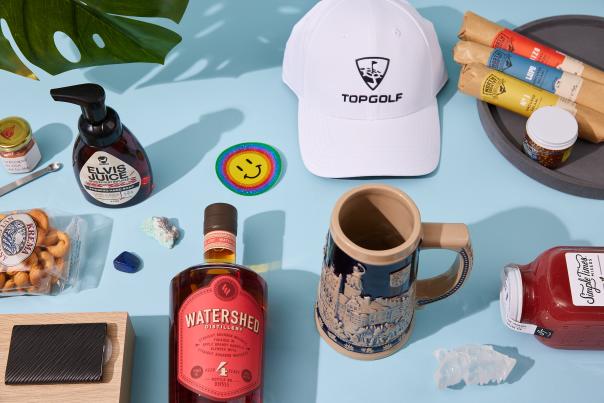 Gift guide items, including a mug, hat, soap, charcuterie items, are artistically arranged on a blue backdrop