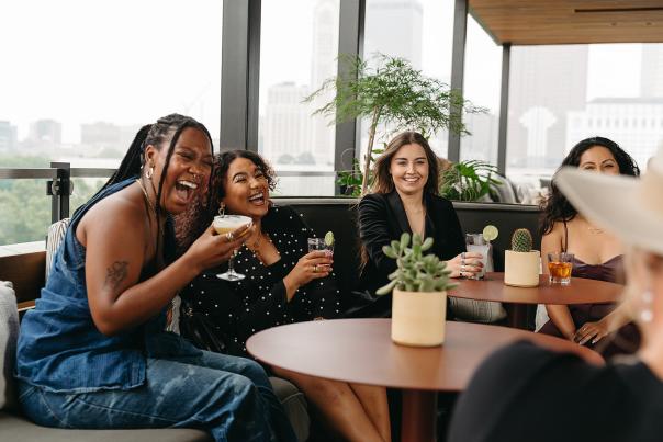 Women sit around a round table while holding cocktails and laughing on a rooftop bar