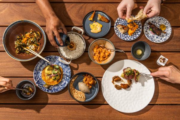 Several plates of food on a wooden table with hands reaching out to pour sauce, grab with forks and pull bread apart