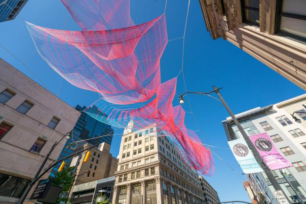 Street view of Janet Echelman's floating scultpture hanging above with urban landscaping in the background