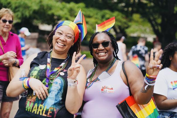 Two women hold up peace signs with their hands and have Pride, rainbow flags in their hair while smiling at the camera