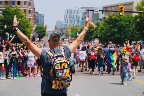 Man with rainbow backpack has arms in the air while walking in PRIDE march