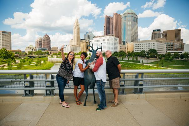 Friends taking picture with deer statue along Scioto Mile. Downtown skyline in the background.