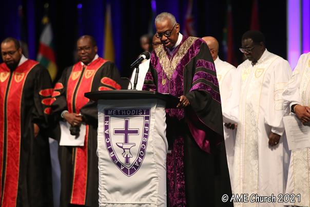 A bishop with the AME Church stands behind a podium with people flanked behind him in different colored robes