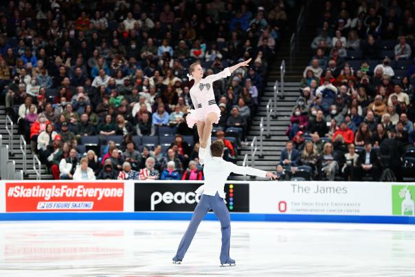 Male figure skater holds female figure skater up with his arm in front of a large arena crowd