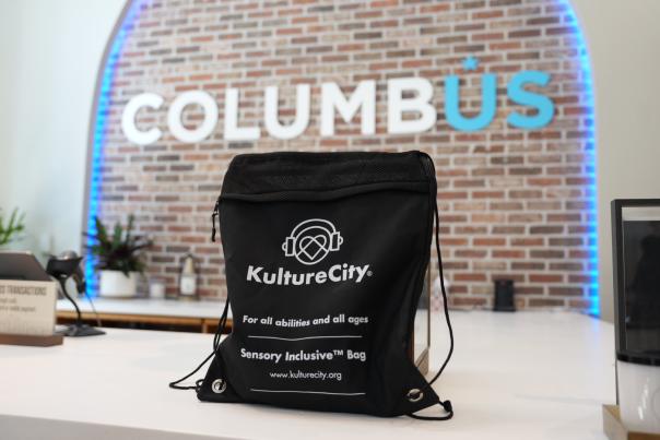 A black KultureCity drawstring sensory bag sits upright on a white counter with a Columbus sign in the background at the Columbus Welcome Center