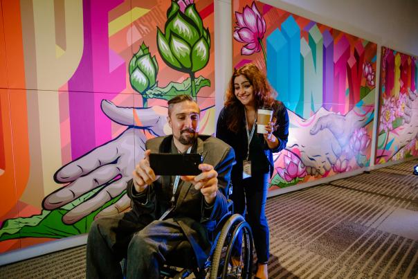 A man and a wheelchair and a woman standing up pose for a selfie in front of a mural at the Greater Columbus Convention Center.