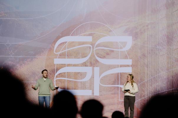 Two people stand onstage beneath a “SEEK” sign, with attendees visible in the foreground.