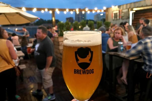 Hand holding full glass of beer with BrewDog's logo, with BrewDog's bustling rooftop patio in background under dark blue sky and twinkle lights